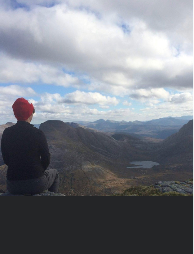 Deb in a red toque sitting on top of the rock overlooking the rolling hills and mountains in the north of Scotland.