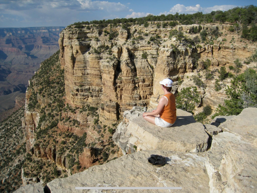 Deb in sukhasana or easy pose overlooking the cliff face of the beautiful grand canyon. Clear blue skies with puffy clouds peak out in the background.